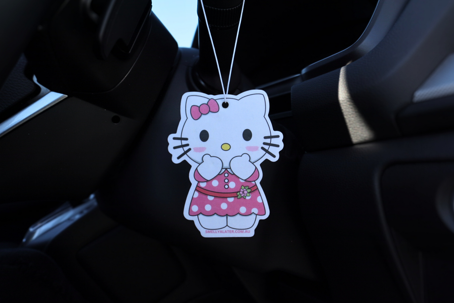Cute blushing Hello Kitty wearing a pink dress with white spots, a pink bow and a flower, hung in a cool black interior car. Smell Ya Later - Scented Air Freshener. Suitable for homes, wardrobes, cupboards, bathrooms, cars, office space, pet rooms, bed rooms, kitchen.