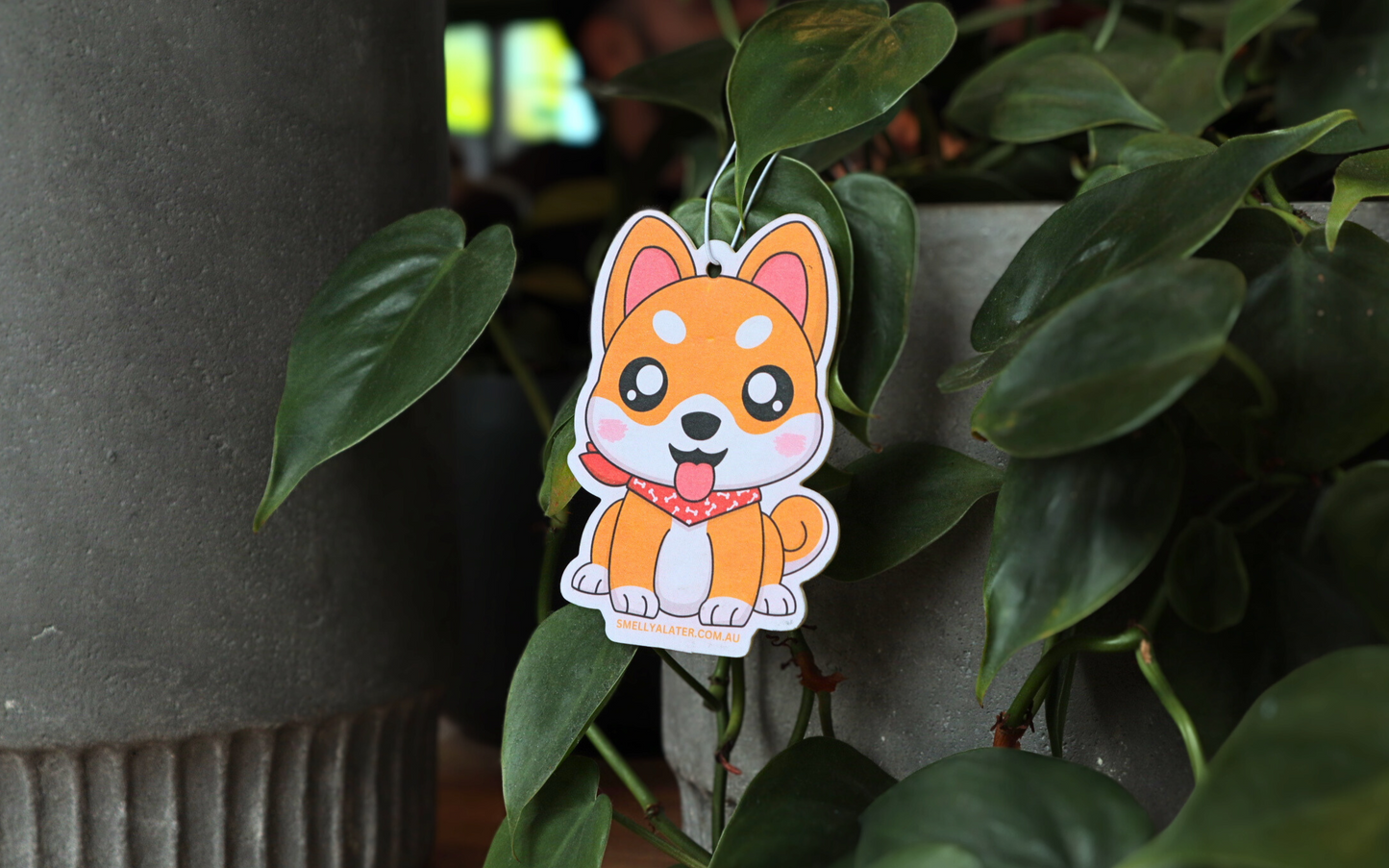 Cute chubby white and orange shiba inu poking its tongue with big sparkling eyes and wearing a red bandanna with white bone patterns, hanging on beautiful green plants. Smell Ya Later - Scented Air Freshener. Suitable for homes, wardrobes, cupboards, bathrooms, cars, office space, pet rooms, bed rooms, kitchen.
