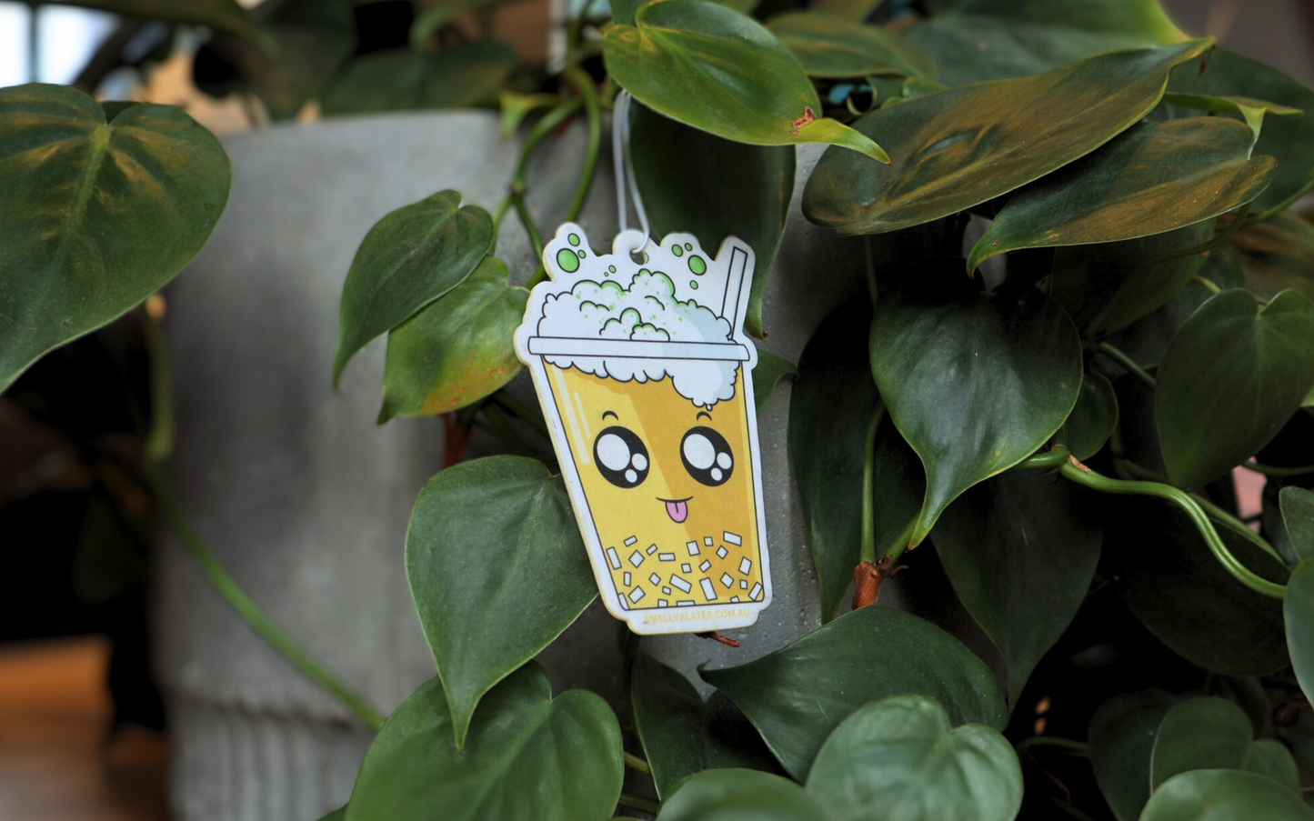 Cute, happy yellow boba/bubble tea poking its tongue with white bubbly froth, green sprinkles, white jelly and big sparkling eyes, hanging on beautiful green plants. Smell Ya Later - Scented Air Freshener. Suitable for homes, wardrobes, cupboards, bathrooms, cars, office space, pet rooms, bed rooms, kitchen.