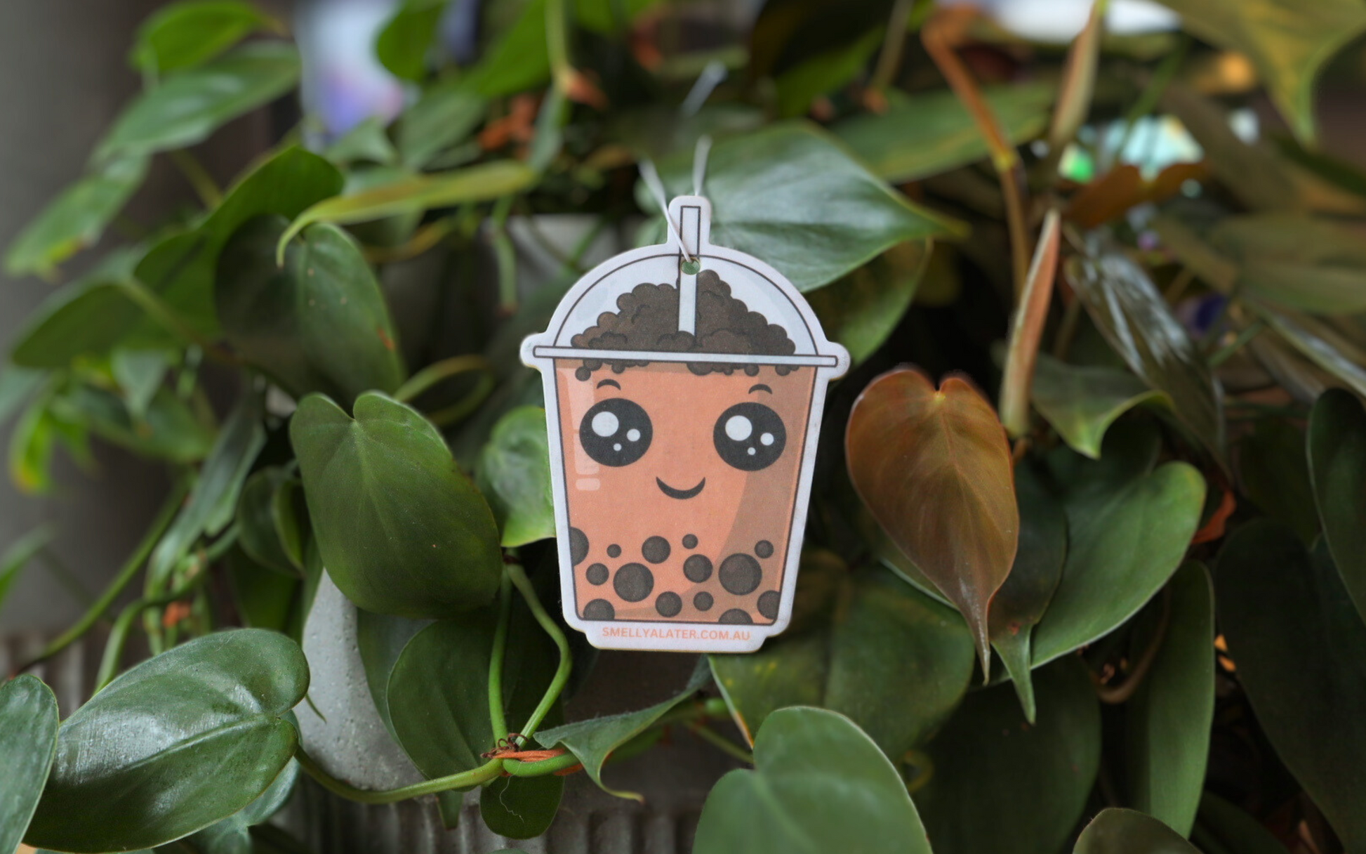 Cute smiling brown boba/bubble tea with black pearls and big sparkling eyes hanging on beautiful green plants. Smell Ya Later - Scented Air Freshener. Suitable for homes, wardrobes, cupboards, bathrooms, cars, office space, pet rooms, bed rooms, kitchen.  